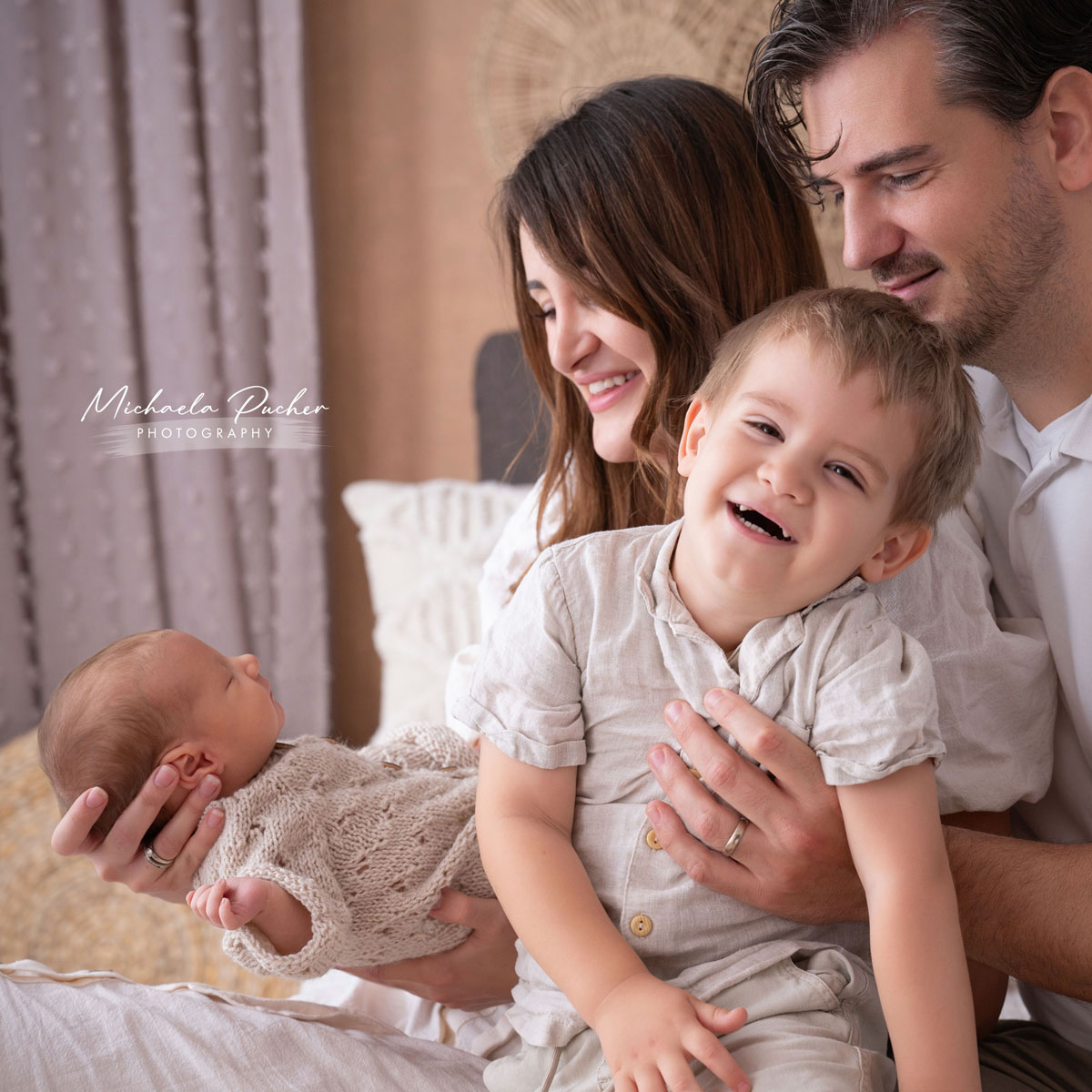 Junge Familie sitzt eng beisammen; der Vater hält das lachende ältere Geschwisterkind, während die Mutter das Neugeborene liebevoll im Arm trägt.